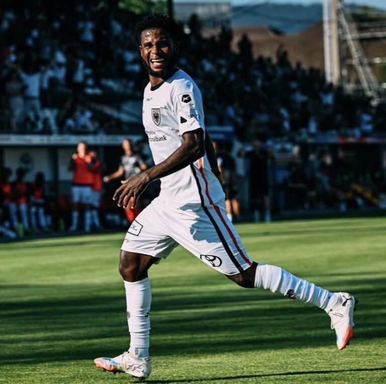 Soccer player in a white kit sprinting on a sunlit green field with a blurred crowd in the stands behind.
