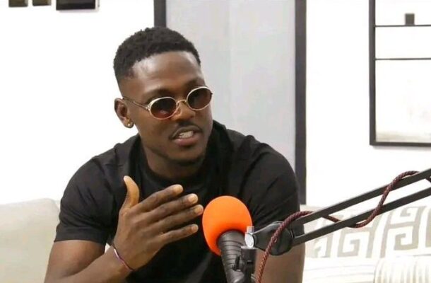 Man wearing round sunglasses speaks into an orange microphone during a podcast session.