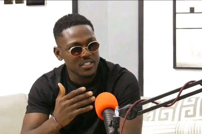 Man wearing round sunglasses speaks into an orange microphone during a podcast session.