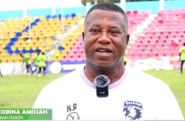 IMG_0375 | Ghanaguardian.com Head coach Kobina Amissah speaks on camera during a football training session; white shirt with crest and lapel mic, colorful stadium seats behind.