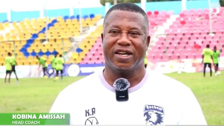 Head coach Kobina Amissah speaks on camera during a football training session; white shirt with crest and lapel mic, colorful stadium seats behind.