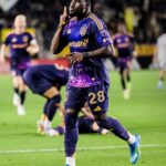 Soccer player in purple LAFC kit (#28) celebrating on the field, pointing to the sky as teammates react in the background (LAFC branding visible).