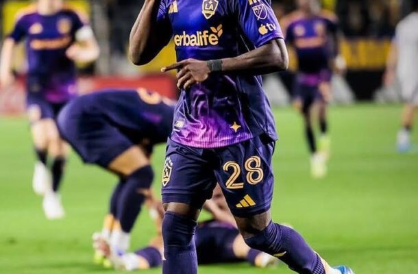 Soccer player in purple LAFC kit (#28) celebrating on the field, pointing to the sky as teammates react in the background (LAFC branding visible).
