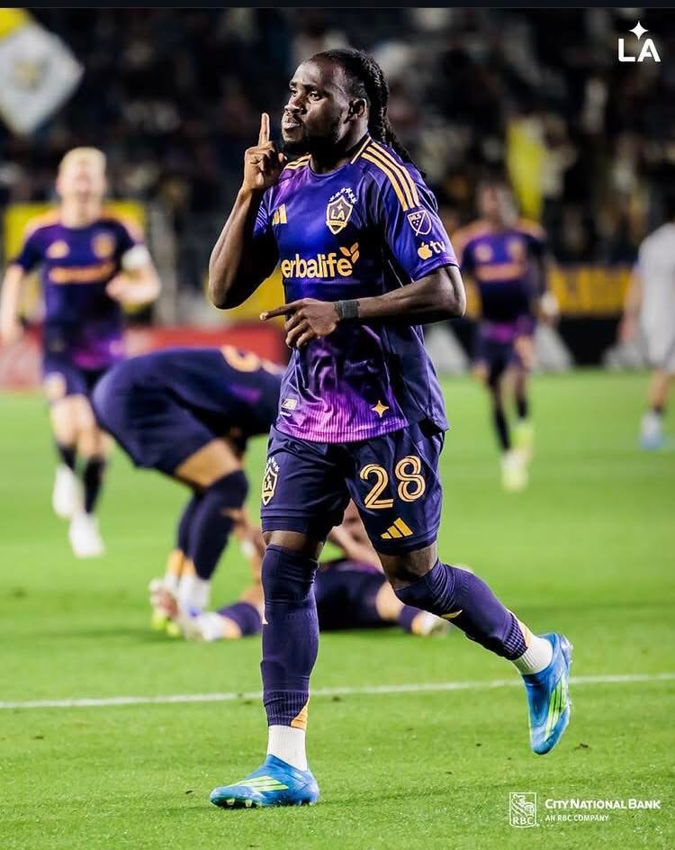 Soccer player in purple LAFC kit (#28) celebrating on the field, pointing to the sky as teammates react in the background (LAFC branding visible).
