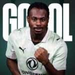 Athlete in a pale green soccer jersey posing with a clenched fist, against a dark green background with the word 'GOAL' in white behind him.