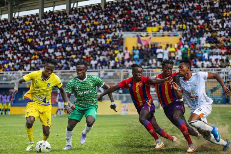 Soccer match scene: players in yellow, green and red-striped kits chase the ball on a grassy field as a large crowd watches from the stands.