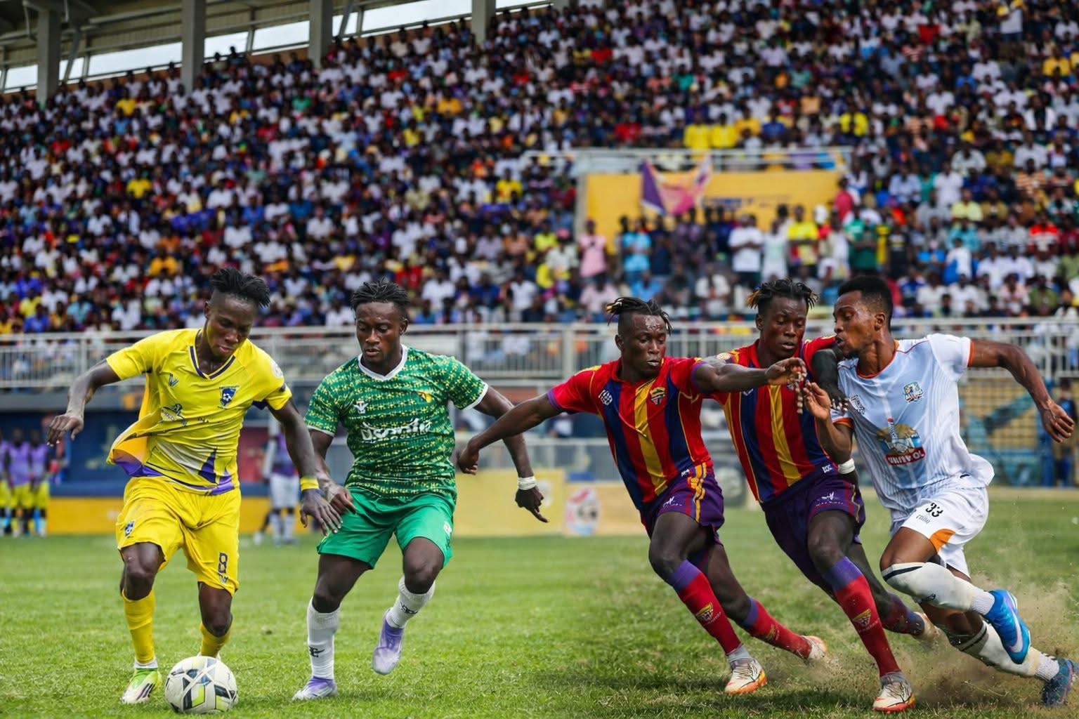 Soccer match scene: players in yellow, green and red-striped kits chase the ball on a grassy field as a large crowd watches from the stands.