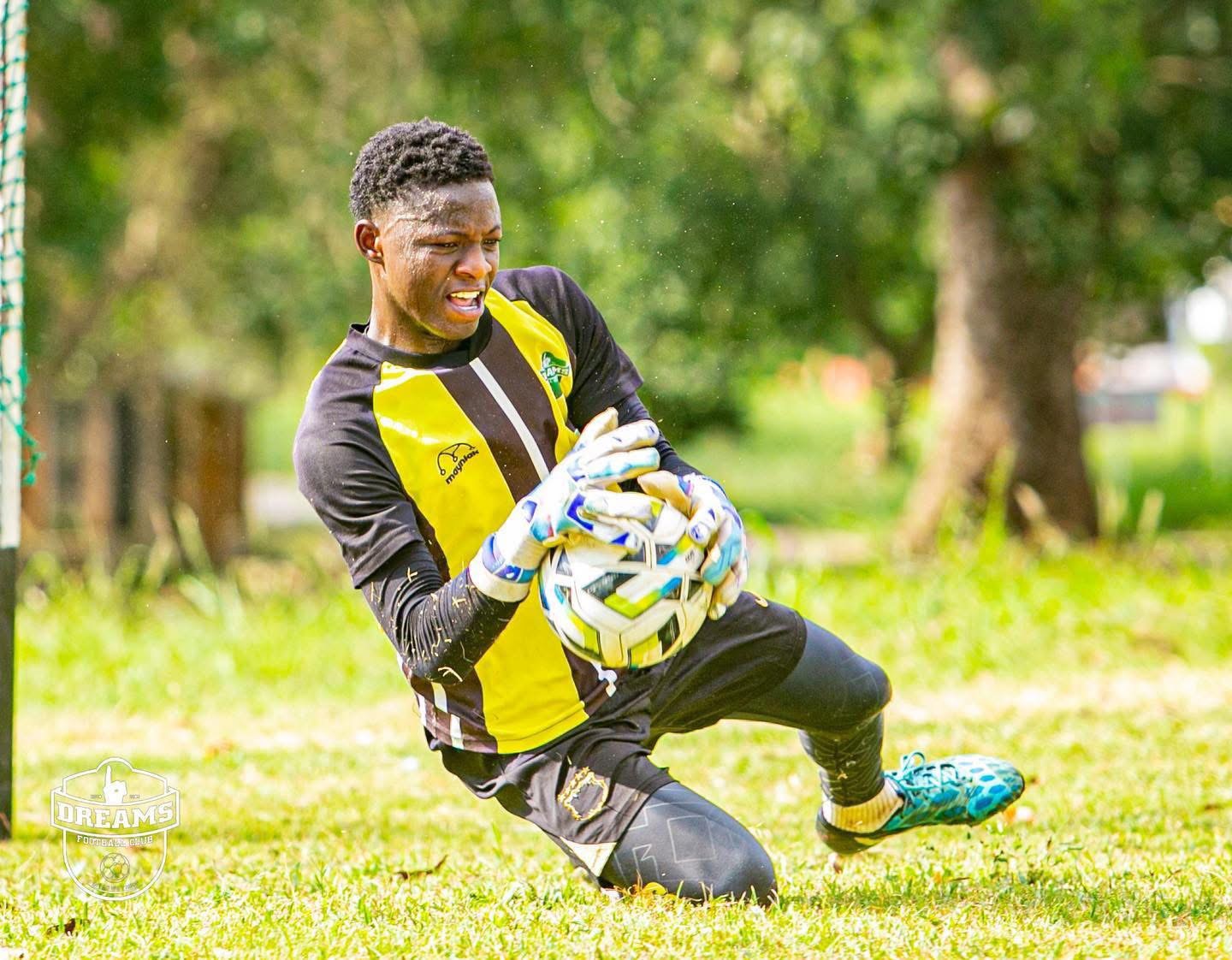 Goalkeeper in yellow and black kit dives to catch a soccer ball on a grassy field with gloves on