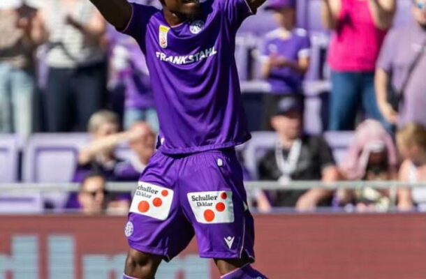 Soccer player in a purple kit raising arms in celebration on the pitch, with fans in the stands behind him.