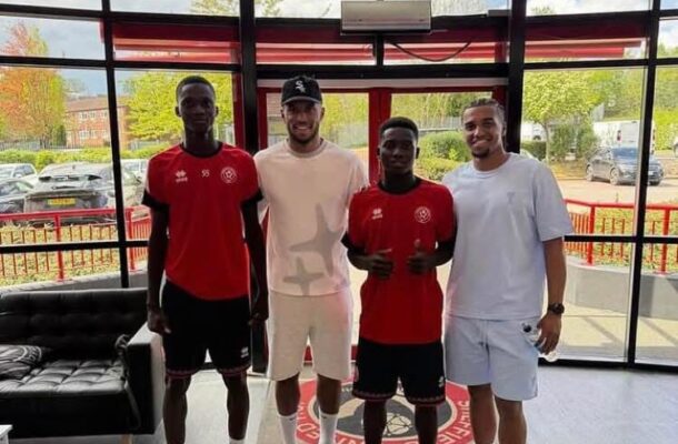 IMG_0413 | Ghanaguardian.com Four men stand side by side indoors in a sports facility, wearing athletic gear (red jerseys and light-colored outfits) and posing for a group photo in front of large glass windows.