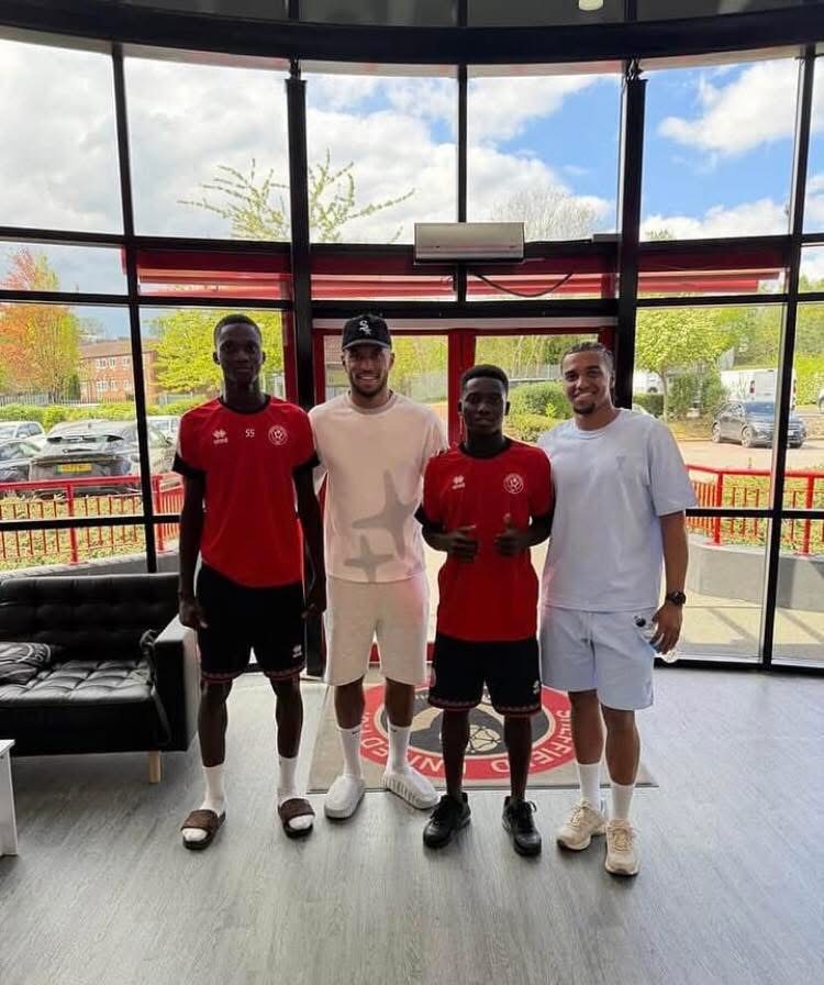 Four men stand side by side indoors in a sports facility, wearing athletic gear (red jerseys and light-colored outfits) and posing for a group photo in front of large glass windows.