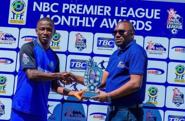 IMG_0422 | Ghanaguardian.com Two men in blue uniforms shake hands while exchanging a trophy at an NBC Premier League Monthly Awards event, with a branded backdrop behind them.