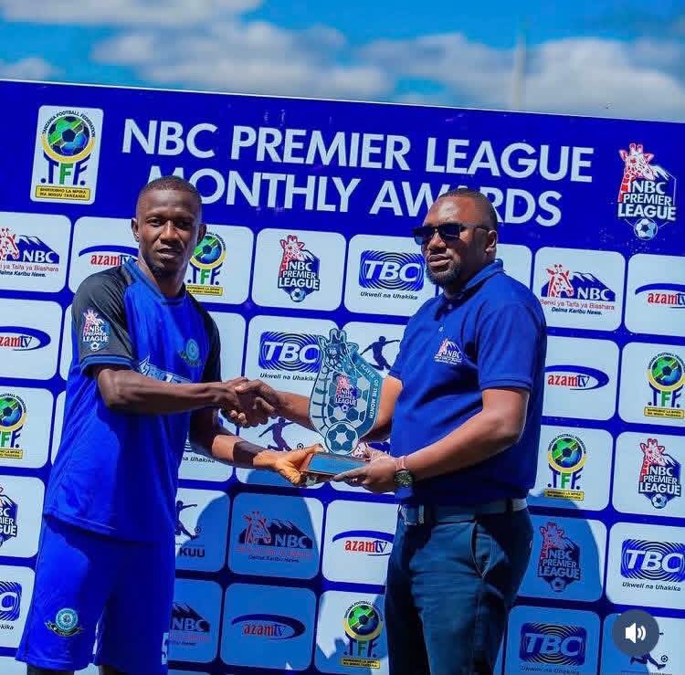 Two men in blue uniforms shake hands while exchanging a trophy at an NBC Premier League Monthly Awards event, with a branded backdrop behind them.