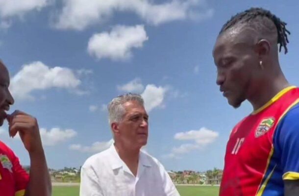 Three men stand on a soccer field in conversation; two players in red jerseys face a man in a white shirt under a blue sky.