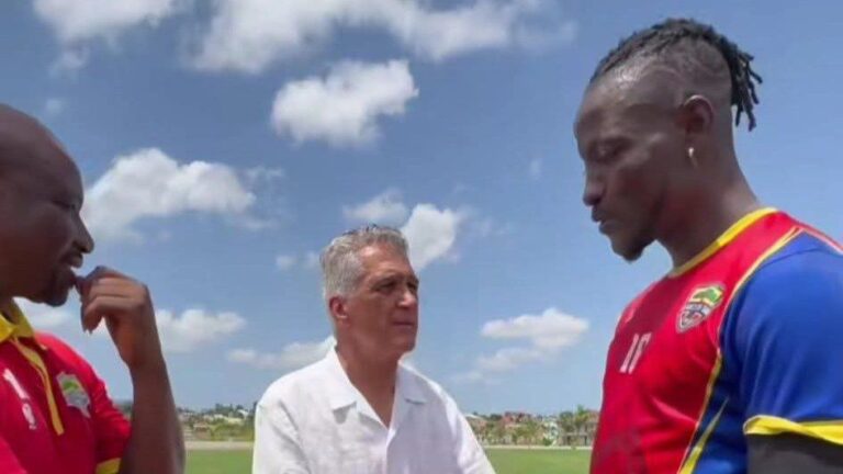 Three men stand on a soccer field in conversation; two players in red jerseys face a man in a white shirt under a blue sky.