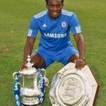 Chelsea footballer in a blue kit kneels on a grass field beside a large silver cup trophy and a shield-shaped trophy, celebrating a win on a soccer pitch.