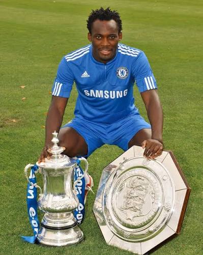 Chelsea footballer in a blue kit kneels on a grass field beside a large silver cup trophy and a shield-shaped trophy, celebrating a win on a soccer pitch.