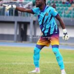 Soccer goalkeeper in turquoise and purple kit, gloves, and teal socks, points to his left on a grassy field during a match.