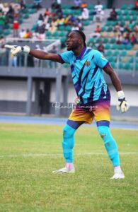 Soccer goalkeeper in turquoise and purple kit, gloves, and teal socks, points to his left on a grassy field during a match.