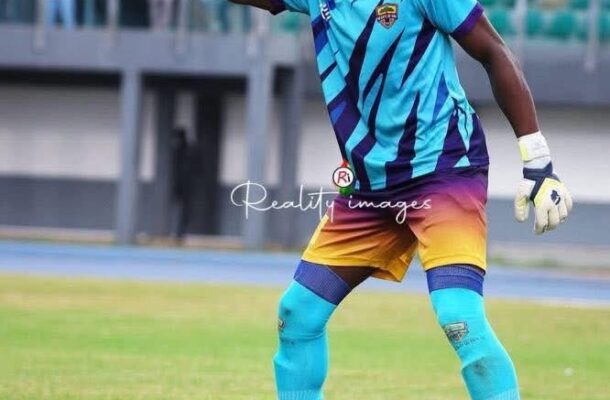 Soccer goalkeeper in turquoise and purple kit, gloves, and teal socks, points to his left on a grassy field during a match.