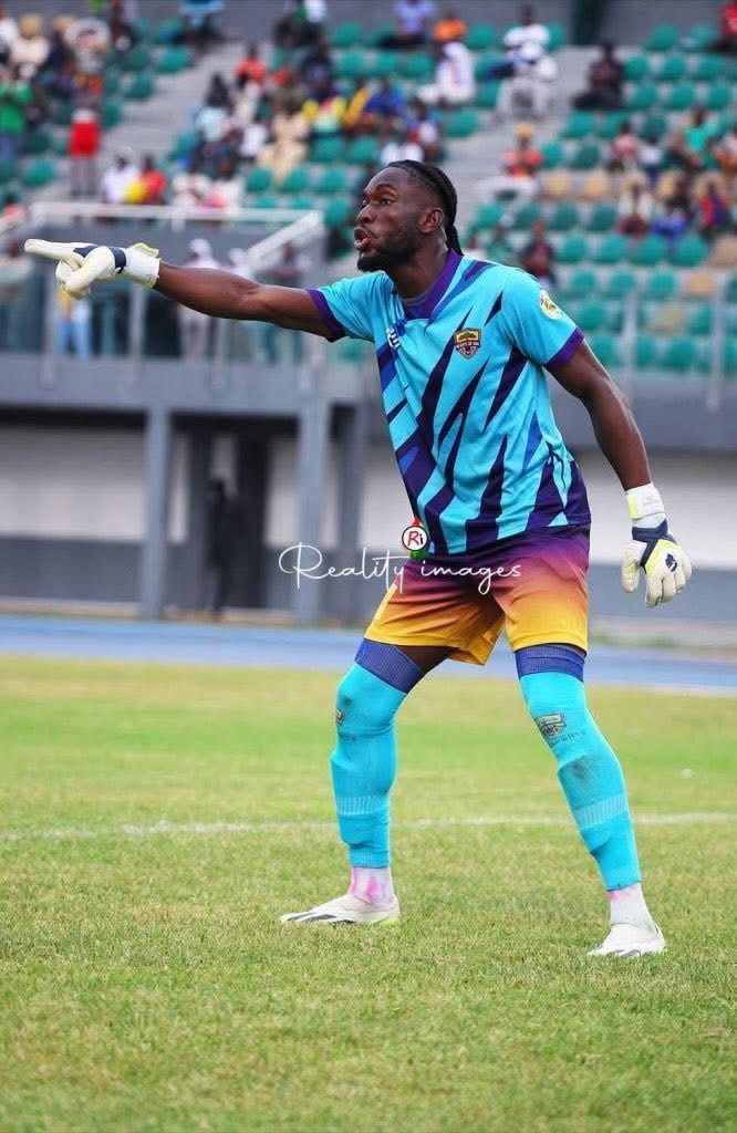 Soccer goalkeeper in turquoise and purple kit, gloves, and teal socks, points to his left on a grassy field during a match.