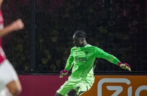 Soccer goalkeeper in a bright green uniform kicking a yellow ball on a night-time pitch with a Ziggo ad in the background.