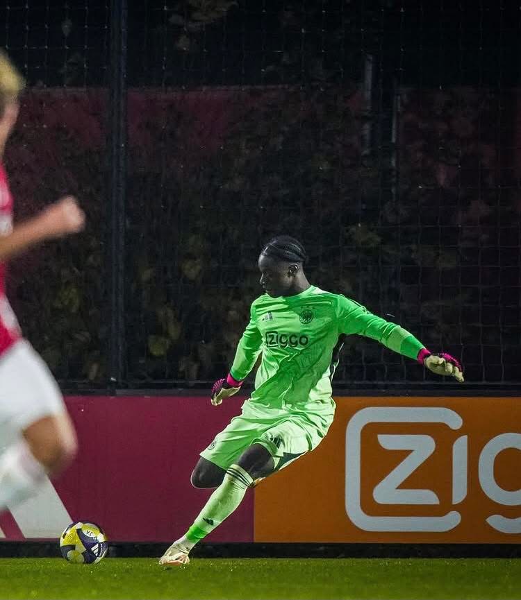 Soccer goalkeeper in a bright green uniform kicking a yellow ball on a night-time pitch with a Ziggo ad in the background.