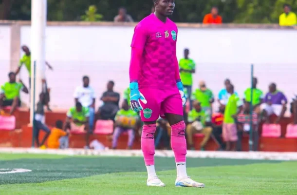 Soccer goalkeeper in a bright pink kit with blue gloves stands on a green pitch as spectators watch in the background.