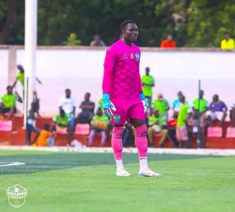 Soccer goalkeeper in a bright pink kit with blue gloves stands on a green pitch as spectators watch in the background.