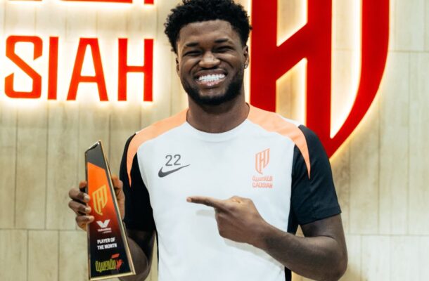 Smiling Black man in a white-orange sports jersey holding a glass 'Player of the Month' trophy, indoors.