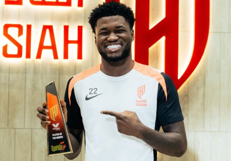 Smiling Black man in a white-orange sports jersey holding a glass 'Player of the Month' trophy, indoors.