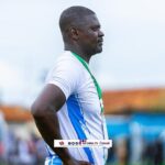 Male athlete in a white and blue jersey, with a green medal lanyard, stands in profile with hands on hips at an outdoor sports event.