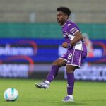 Soccer player in a purple kit kicking a ball on a rain-soaked field during a match without a visible crowd in the background.— Informative action description