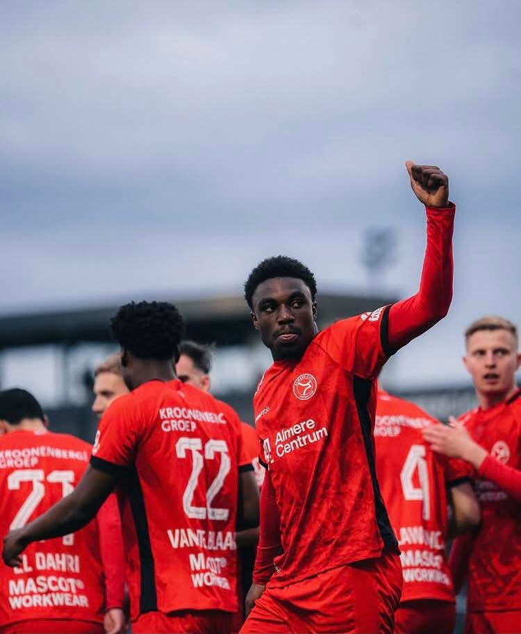 Soccer players in red jerseys celebrate on the field, the central player raising his right arm in a fist.