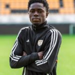 Young Black male soccer player standing on a green field with arms crossed, wearing a black training jacket with white stripes and an SJK crest.