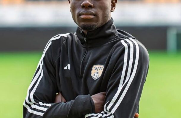 Young Black male soccer player standing on a green field with arms crossed, wearing a black training jacket with white stripes and an SJK crest.