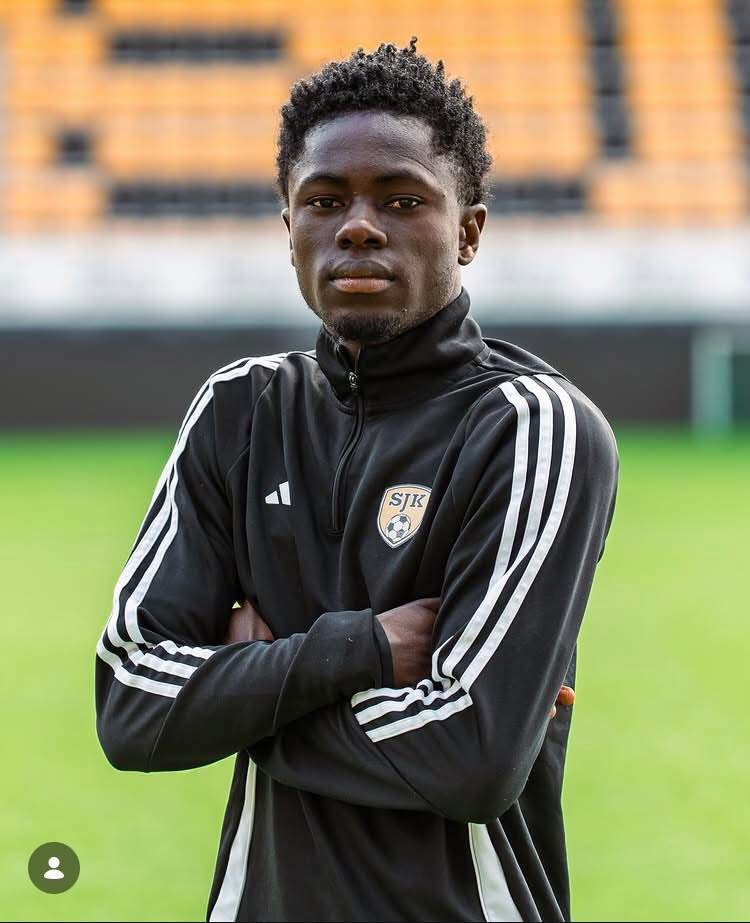 Young Black male soccer player standing on a green field with arms crossed, wearing a black training jacket with white stripes and an SJK crest.