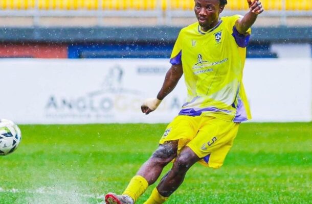 Soccer player in a bright yellow kit kicking a ball on a wet, grassy field in a match.