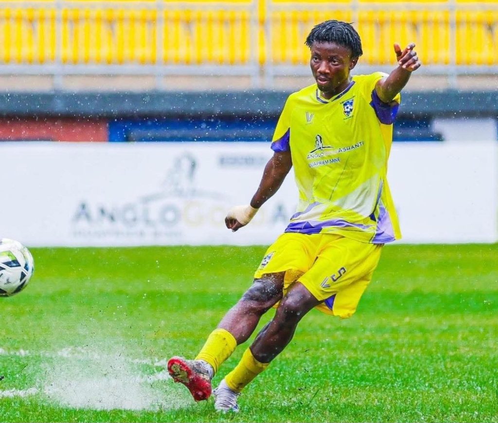 Soccer player in a bright yellow kit kicking a ball on a wet, grassy field in a match.