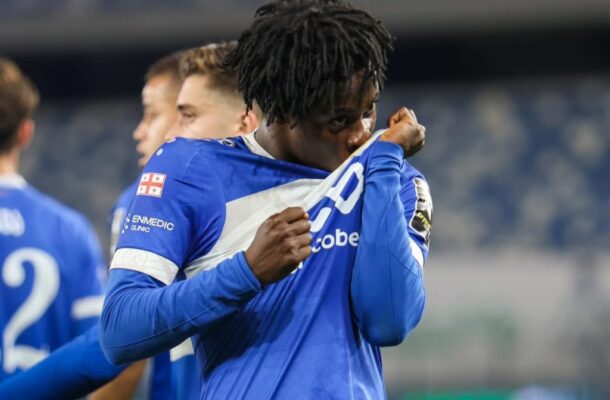 Soccer player in a blue kit celebrates by pulling his jersey over his face on the field, teammates blurred in the background.