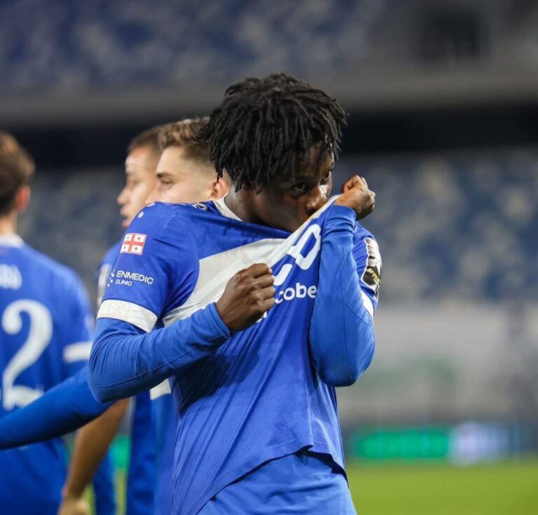 Soccer player in a blue kit celebrates by pulling his jersey over his face on the field, teammates blurred in the background.