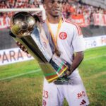 Soccer player in a white kit holds a tall trophy with a gold medal around his neck, stadium crowd in the background.