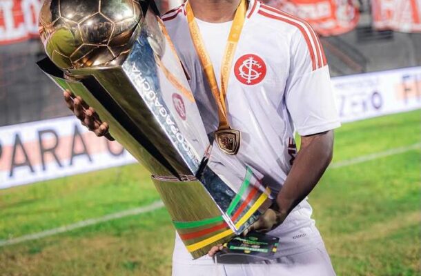 Soccer player in a white kit holds a tall trophy with a gold medal around his neck, stadium crowd in the background.