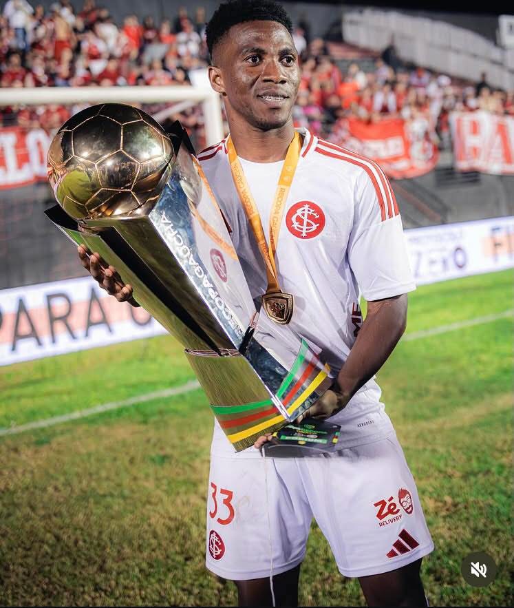 Soccer player in a white kit holds a tall trophy with a gold medal around his neck, stadium crowd in the background.