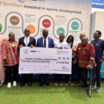 Group of eight people posing with a large ceremonial check for five million dollars to the Ghana Football Association, standing in front of a display about sports development and investment missives on the backdrop.