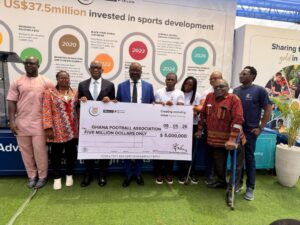 Group of eight people posing with a large ceremonial check for five million dollars to the Ghana Football Association, standing in front of a display about sports development and investment missives on the backdrop.