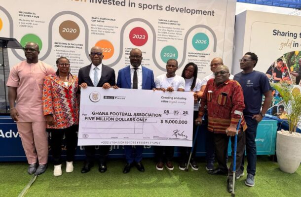 Group of eight people posing with a large ceremonial check for five million dollars to the Ghana Football Association, standing in front of a display about sports development and investment missives on the backdrop.