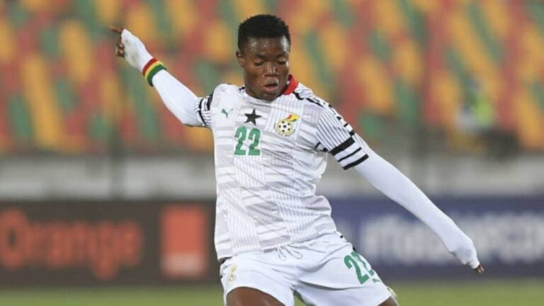 Soccer player in a white kit with number 22 mid-kick on a stadium field with colorful empty stands behind him.