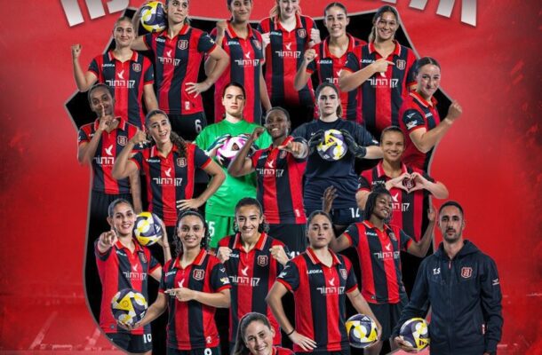 Group portrait of a women's volleyball team in red-and-black jerseys posing together; several players hold volleyballs, with a coach on the right, set against a red background with large Hebrew text that reads 'State Champion'.