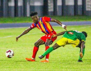 Soccer players contesting the ball on a green field; one in a red-yellow striped jersey and red shorts, the other in green and yellow.